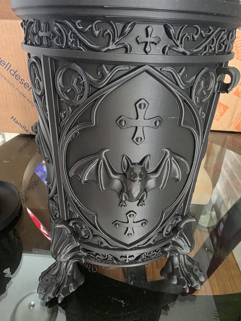 Close-up of gothic bat canister showing ornate carved details, raised bat emblem, cross accents, and clawed feet, highlighting dark gothic craftsmanship and textured matte black finish.