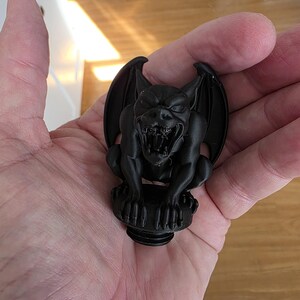 Close-up of a black gothic gargoyle topper held in a hand, showing a snarling expression with open mouth, sharp teeth, folded wings, clawed feet, and a matte finish, designed to screw into the lid of a gothic decorative canister.