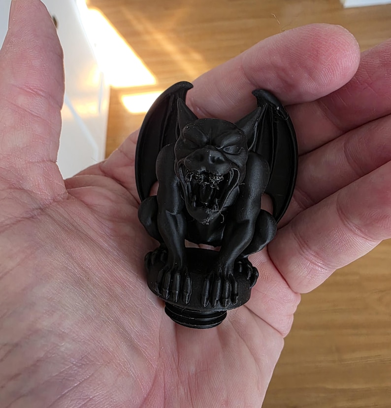 Close-up of a black gothic gargoyle topper held in a hand, showing a snarling expression with open mouth, sharp teeth, folded wings, clawed feet, and a matte finish, designed to screw into the lid of a gothic decorative canister.
