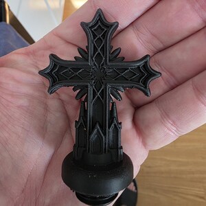 Close-up of a black gothic cross topper resting in a hand, featuring ornate layered details, sharp architectural lines, and a matte finish, designed to screw into the lid of a gothic decorative storage canister.
