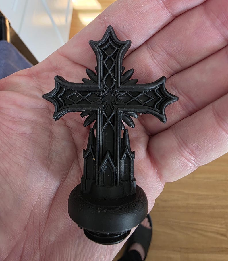 Close-up of a black gothic cross topper resting in a hand, featuring ornate layered details, sharp architectural lines, and a matte finish, designed to screw into the lid of a gothic decorative storage canister.