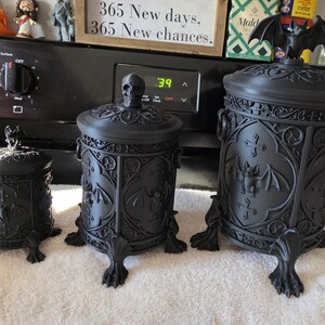 Three gothic storage canisters displayed side by side in small, medium, and large sizes, featuring ornate gothic detailing and bat-themed designs, shown as dark home decor organizer jars for bathroom, kitchen, or office use.
