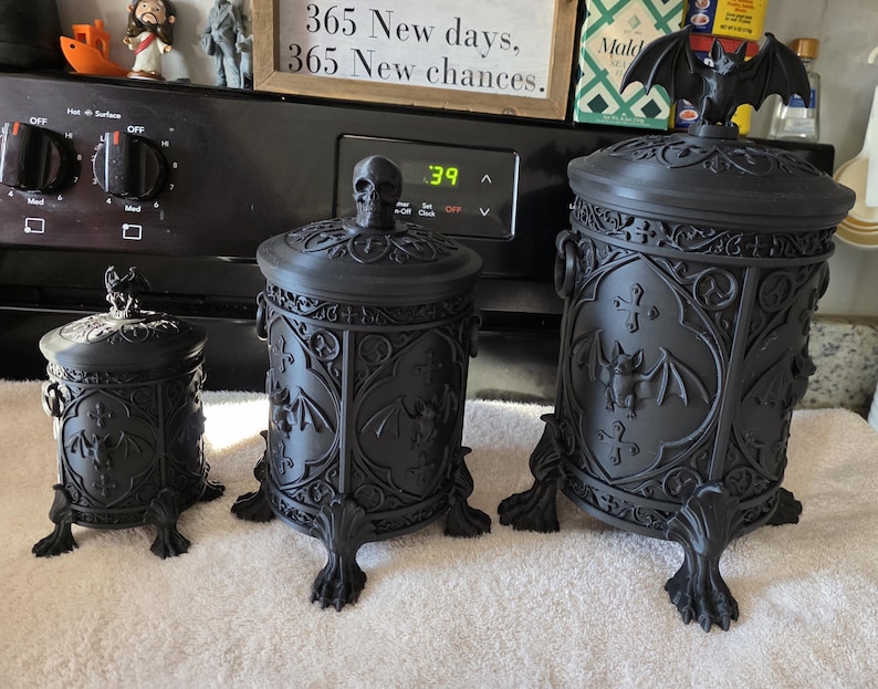 Three gothic storage canisters displayed side by side in small, medium, and large sizes, featuring ornate gothic detailing and bat-themed designs, shown as dark home decor organizer jars for bathroom, kitchen, or office use.