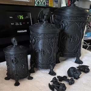 Three gothic bat canisters displayed together in small, medium, and large sizes, featuring ornate carved details, bat emblems, clawed feet, and matching lids, with interchangeable bat and skull toppers shown in front to demonstrate size and style options.