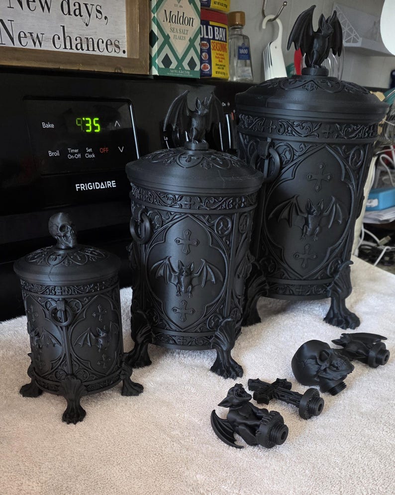 Three gothic bat canisters displayed together in small, medium, and large sizes, featuring ornate carved details, bat emblems, clawed feet, and matching lids, with interchangeable bat and skull toppers shown in front to demonstrate size and style options.