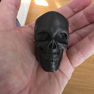 Close-up of a black gothic skull topper resting in a hand, showing detailed facial features, smooth matte finish, and a compact sculpted design intended to attach to the lid of a gothic storage canister.