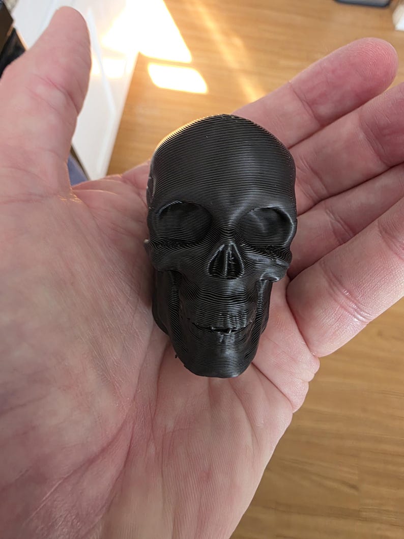 Close-up of a black gothic skull topper resting in a hand, showing detailed facial features, smooth matte finish, and a compact sculpted design intended to attach to the lid of a gothic storage canister.