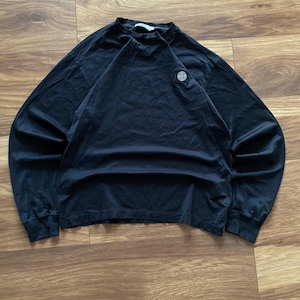 Puede incluir: Una camiseta negra de manga larga con cuello redondo. Un pequeño emblema circular está en el pecho izquierdo. La camiseta está hecha de un material suave y está colocada sobre una superficie de madera. Las mangas son largas y llegan a las muñecas.