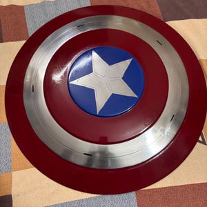 May include: A circular shield featuring a glossy dark red outer ring, a metallic silver middle ring, and a vibrant blue centre circle. A large, five-pointed silver star is centred within the blue circle, displaying subtle geometric line details.