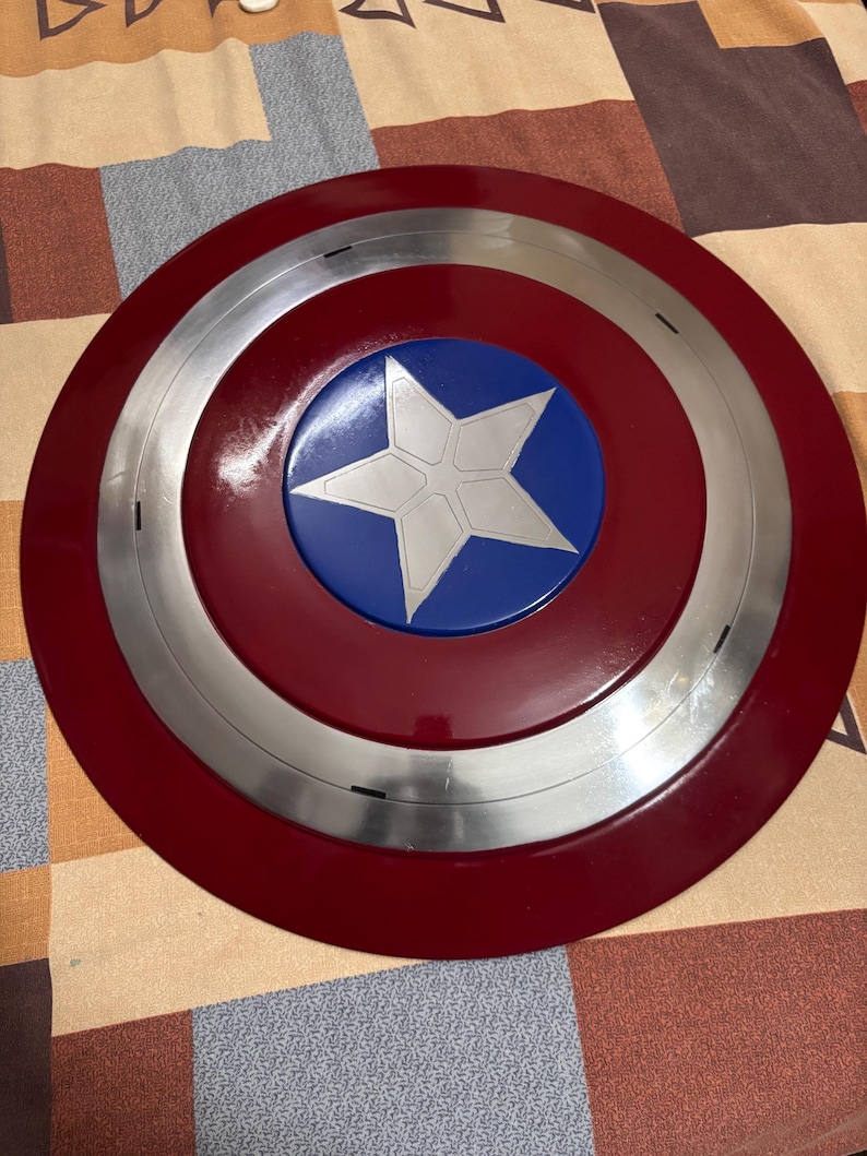 May include: A circular shield featuring a glossy dark red outer ring, a metallic silver middle ring, and a vibrant blue centre circle. A large, five-pointed silver star is centred within the blue circle, displaying subtle geometric line details.