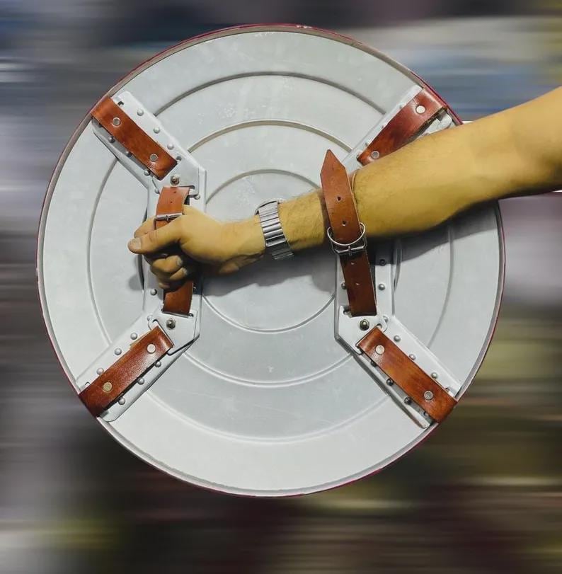 May include: A silver and red shield with brown leather straps and rivets. The shield is round with a silver center and a red border. The straps are attached to the back of the shield.