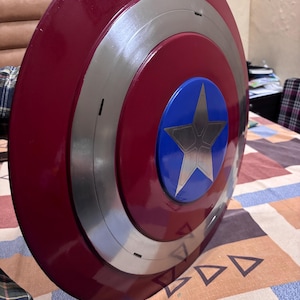 May include: A circular shield featuring a glossy deep red outer ring, a metallic silver middle ring, and a bright blue centre circle. A large, five-pointed silver star is centred within the blue circle, creating a bold, multi-layered design.