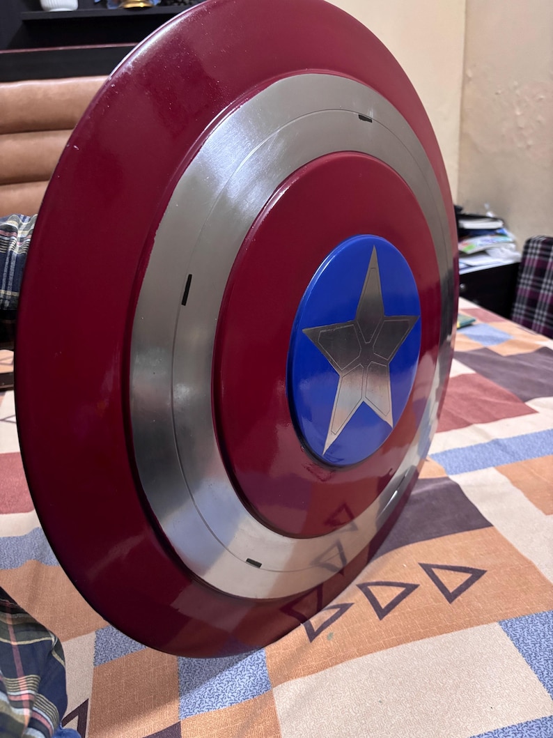 May include: A circular shield featuring a glossy deep red outer ring, a metallic silver middle ring, and a bright blue centre circle. A large, five-pointed silver star is centred within the blue circle, creating a bold, multi-layered design.