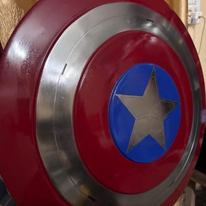 May include: A circular shield featuring a glossy red outer ring, a metallic silver middle ring, and a vibrant blue centre circle. A large, five-pointed silver star is centred within the blue circle, creating a bold, multi-layered design.
