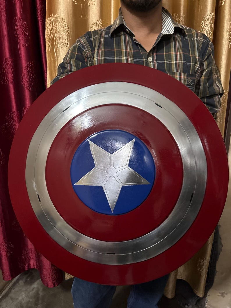 May include: A circular shield featuring a glossy red outer ring, a metallic silver middle ring, and a central blue circle with a large, silver five-pointed star. The design is symmetrical, with clean lines and a polished finish throughout the surface.
