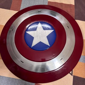 May include: A circular shield featuring a deep red outer ring, a metallic silver middle ring, and a vibrant blue centre circle. A large, five-pointed white star is centred within the blue circle, displaying subtle etched lines across its surface.