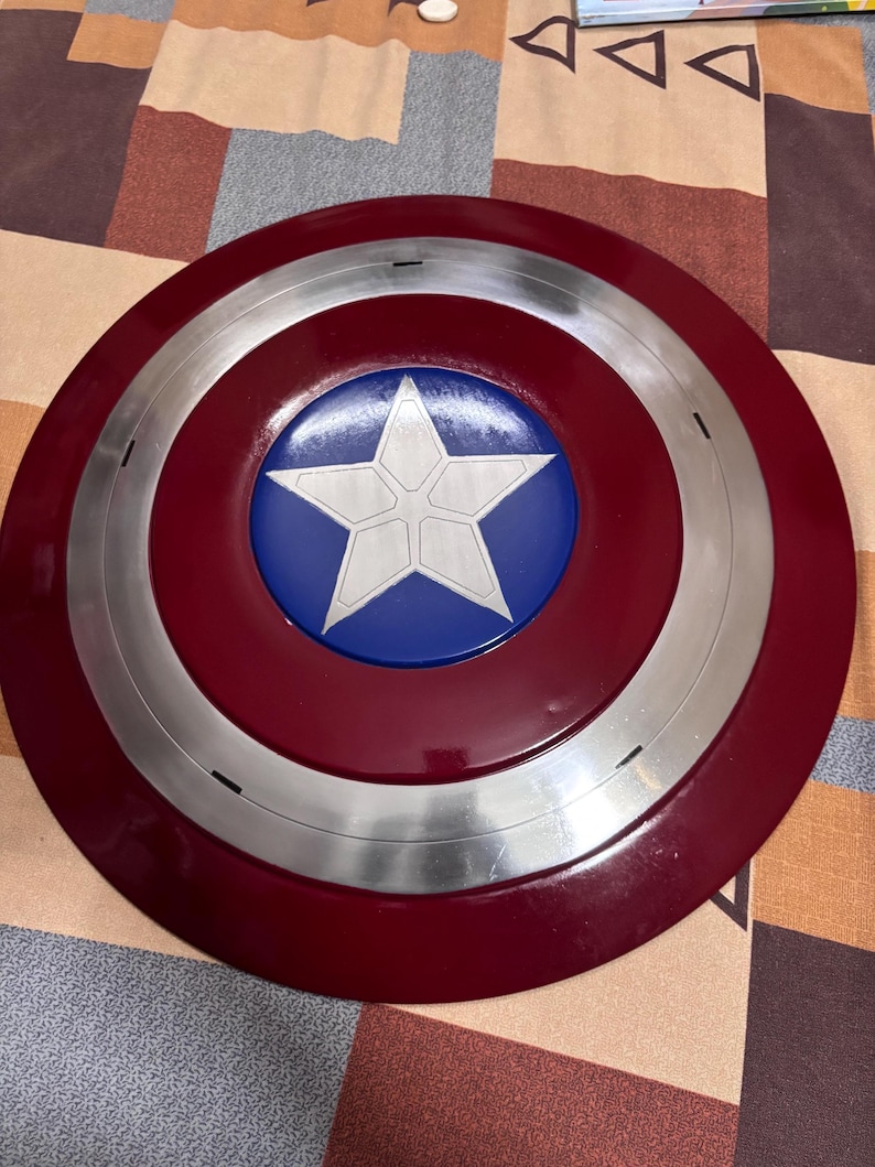 May include: A circular shield featuring a deep red outer ring, a metallic silver middle ring, and a vibrant blue centre circle. A large, five-pointed white star is centred within the blue circle, displaying subtle etched lines across its surface.