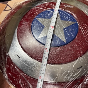May include: A circular shield featuring a central silver star on a blue background, surrounded by alternating red and silver rings. A measuring tape placed across the diameter shows a measurement of approximately 58 centimetres in total width.