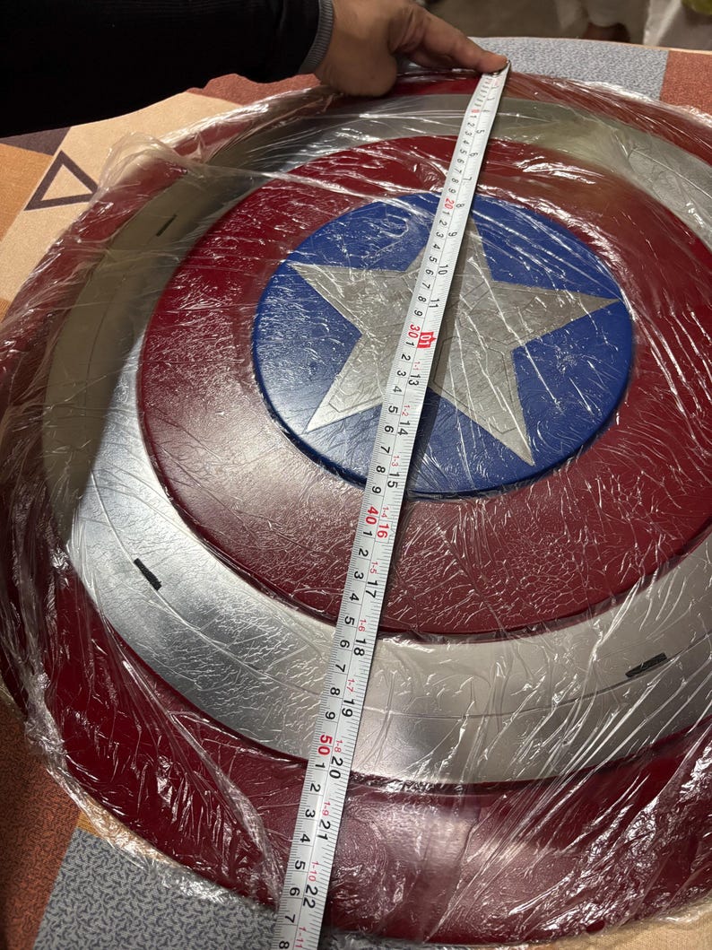 May include: A circular shield featuring a central silver star on a blue background, surrounded by alternating red and silver rings. A measuring tape placed across the diameter shows a measurement of approximately 58 centimetres in total width.