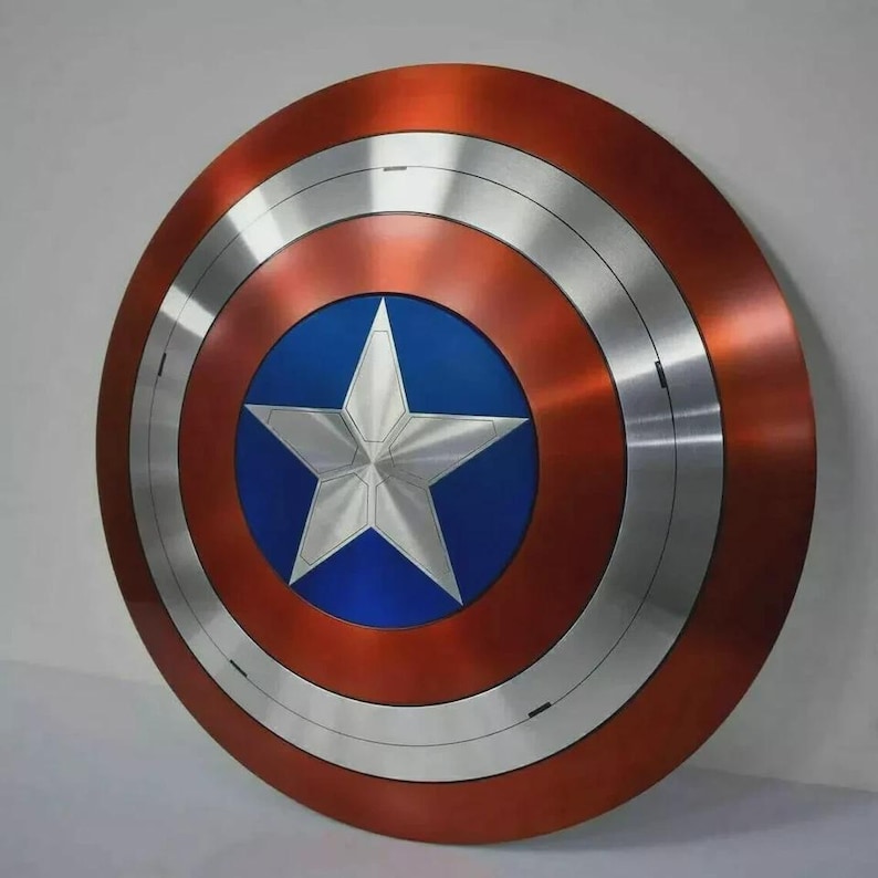 May include: A round shield with a red, silver, and blue color scheme. The shield features a silver star in the center, surrounded by a blue circle. The outer ring is red, with silver bands separating the colors. A superhero-themed item.
