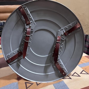 May include: A circular grey metal shield featuring a smooth, concentric ring design. The back side includes two sets of brown leather straps with silver-toned metal buckles and rivets, secured to the shield by grey metal mounting brackets.