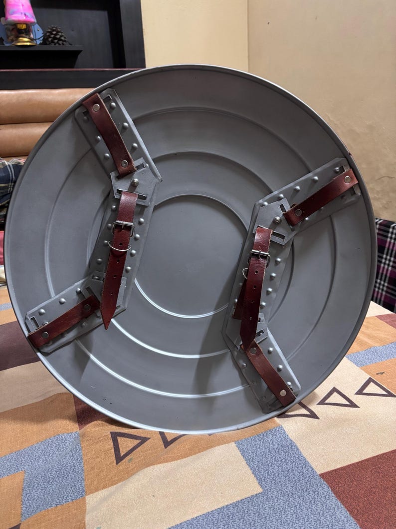 May include: A circular grey metal shield featuring a smooth, concentric ring design. The back side includes two sets of brown leather straps with silver-toned metal buckles and rivets, secured to the shield by grey metal mounting brackets.