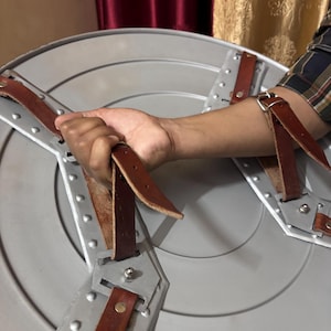May include: A circular silver-coloured shield featuring a metallic surface with concentric circular ridges. The interior side includes two brown leather straps secured by metal rivets and brackets, designed for forearm and hand attachment.