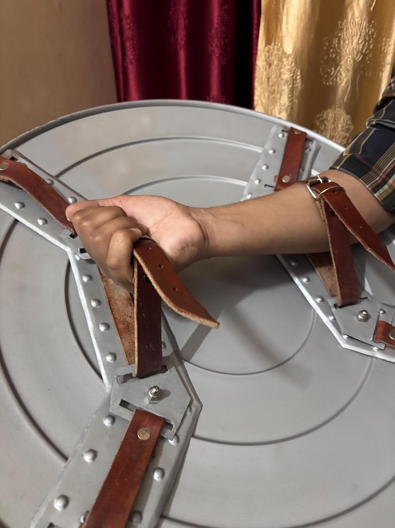 May include: A circular silver-coloured shield featuring a metallic surface with concentric circular ridges. The interior side includes two brown leather straps secured by metal rivets and brackets, designed for forearm and hand attachment.