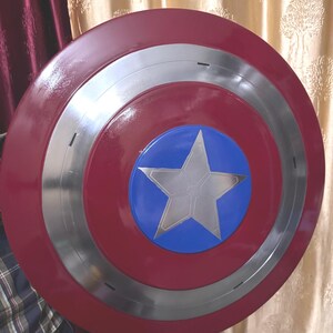 May include: A circular shield featuring a glossy red outer ring, a metallic silver middle ring, and a central blue circle containing a large, five-pointed silver star. The design is symmetrical with a smooth, reflective finish across the surface.