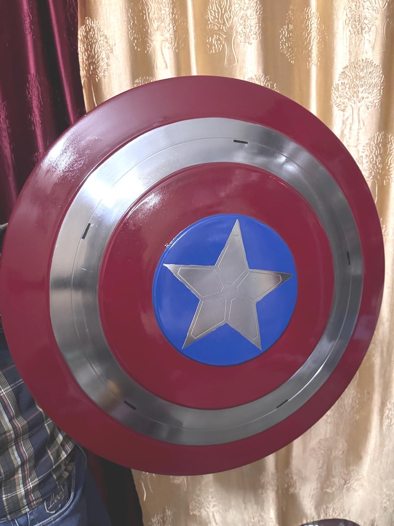 May include: A circular shield featuring a glossy red outer ring, a metallic silver middle ring, and a central blue circle containing a large, five-pointed silver star. The design is symmetrical with a smooth, reflective finish across the surface.