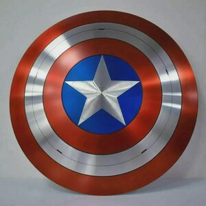 May include: A circular shield with a red outer ring, a silver band, and a blue center. A large, five-pointed silver star is centered on the blue circle. The shield has a metallic appearance and is likely a prop or collectible.