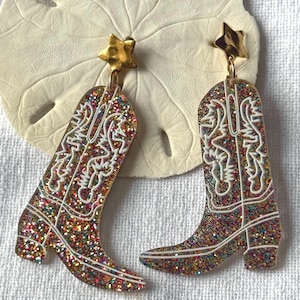 Könnte beinhalten: Ein Paar glitzernde Cowboy-Stiefel-Ohrringe. Die Ohrringe haben ein farbenfrohes, gesprenkeltes Design mit weißen Details und einem goldenen Stern oben. Die Stiefel haben ein klassisches Western-Design.