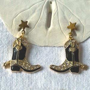 Könnte beinhalten: Goldfarbene Ohrringe mit schwarzen Cowboy-Stiefel-Anhängern mit Strasssteinen. Jeder Stiefel hat ein goldenes Herz und eine Spore und ist an einem goldfarbenen sternförmigen Stecker befestigt.