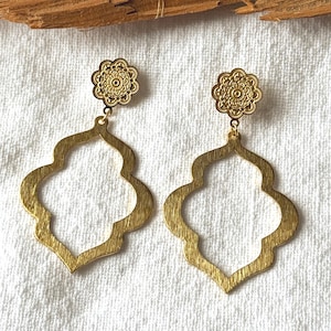 Op de afbeelding: Een paar goudkleurige oorhangers. Elke oorbel heeft een bloemenstud en een grote, open vierpasvorm. De oorbellen worden gepresenteerd op een wit oppervlak, met een stuk hout op de achtergrond. De oorbellen zijn een sieraad.