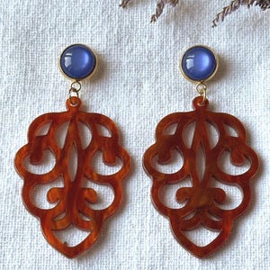 Op de afbeelding: Een paar oorbellen met een blauwe cabochon-top met een goudkleurige zetting, verbonden met een decoratieve, bladvormige hanger. De hanger heeft een warme, roodbruine kleur met een ingewikkeld, opengewerkt ontwerp. Op een witte achtergrond.