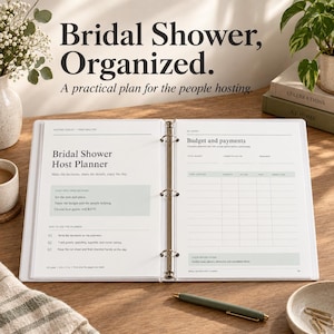 May include: A white three-ring binder open to pages titled Bridal Shower Host Planner and Budget and payments. The pages feature sections for planning decisions, guest lists, and financial tracking. A green pen lies on the wooden surface nearby.