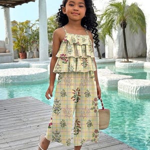 May include: A yellow plaid two-piece outfit with floral accents. The top has a ruffled design, and the pants are wide-legged. The outfit is paired with white sandals and a small woven handbag. The outfit is designed for children.