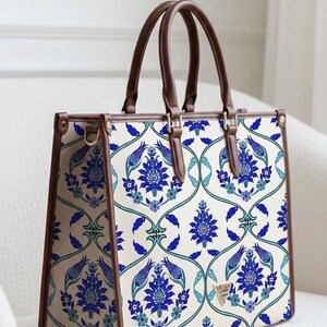 May include: A rectangular tote bag with a cream background and a blue floral pattern. The bag has brown handles and trim, with gold-colored hardware. The bag is sitting on a white surface.