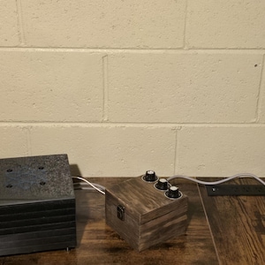 May include: Two wooden electronic devices with knobs and a black metal box are connected by white wires on a wooden surface. The background is a light beige brick wall with a floral design.