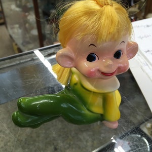 May include: A yellow-haired doll with green pants and a yellow shirt. The doll has a smiling face with blue eyes and is sitting with its legs crossed.