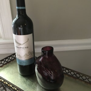 Op de afbeelding: Een fles 2018 Trapiche Malbec wijn uit Argentini&euml;, met een donkerpaarse glazen vaas op een zilveren dienblad met een zwart metalen frame.