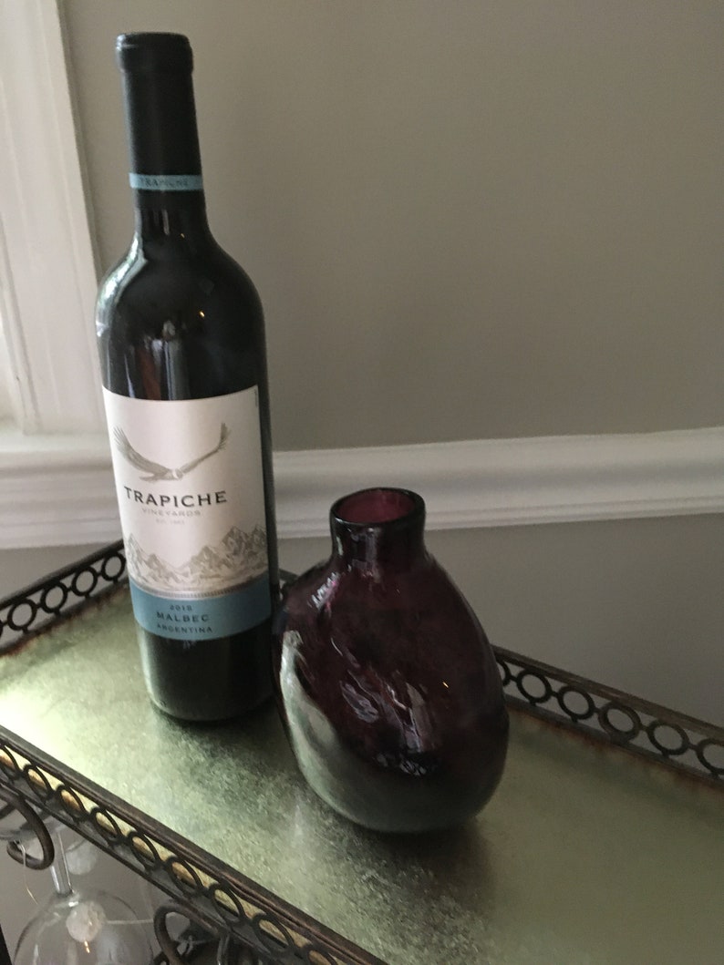 Op de afbeelding: Een fles 2018 Trapiche Malbec wijn uit Argentini&euml;, met een donkerpaarse glazen vaas op een zilveren dienblad met een zwart metalen frame.