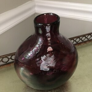 Op de afbeelding: Een ronde, paarse glazen vaas met een lichte iriserende glans. De vaas heeft een brede opening en een smalle hals.