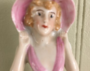 May include: A porcelain figurine of a woman wearing a pink hat and dress. The figurine has a delicate, vintage style.