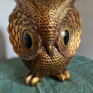 May include: A gold owl figurine with large, round eyes. The owl has a textured surface and is standing on a green surface.