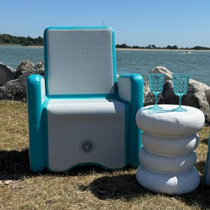 Può includere: Una sedia gonfiabile da esterno con seduta bianca e struttura turchese è appoggiata sull'erba vicino all'acqua. Un tavolino laterale in pietra bianca impilata contiene due bicchieri blu a fantasia. La scena è ambientata in una giornata di sole.