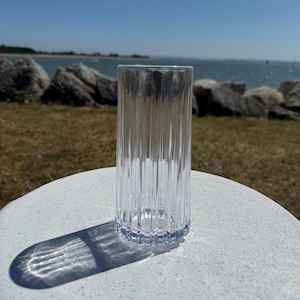 Puede incluir: Un vaso de vidrio transparente y acanalado se encuentra sobre una superficie blanca, proyectando una sombra. El vaso es cilíndrico con estrías verticales. El fondo presenta una zona de césped, rocas y el océano bajo un cielo azul brillante.