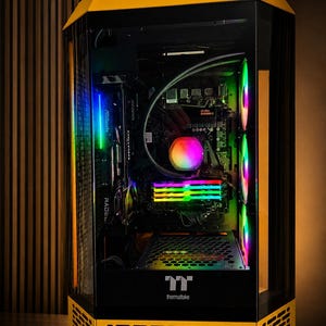 May include: A yellow and black Thermaltake computer tower case with a transparent side panel. The interior features a motherboard with ASRock branding, multi-colored RGB lighting on the cooling fans, RAM sticks, and a central processor liquid cooler.