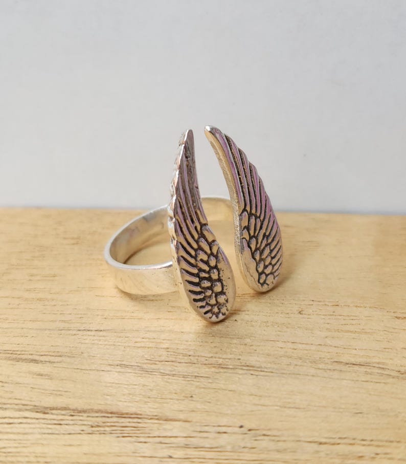 May include: A silver-colored ring featuring an open design with two detailed wing shapes. The wings are positioned to appear as if they are wrapping around a finger. The ring is set against a light wood surface.