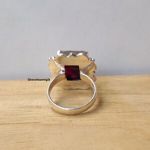 Pu&ograve; includere: Un anello d'argento con una grande gemma quadrata di colore rosso scuro al centro. La gemma &egrave; circondata da un'incastonatura decorativa e strutturata. L'anello &egrave; appoggiato su una superficie di legno. Il testo "Silverartisanring26" &egrave; visibile.